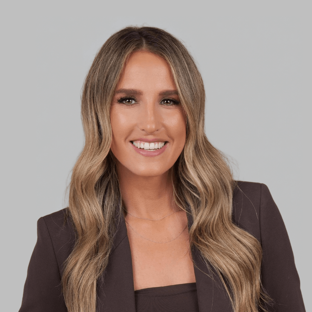 Smiling woman with long, wavy light brown hair wearing a dark blazer and top, posing against a plain light gray background.