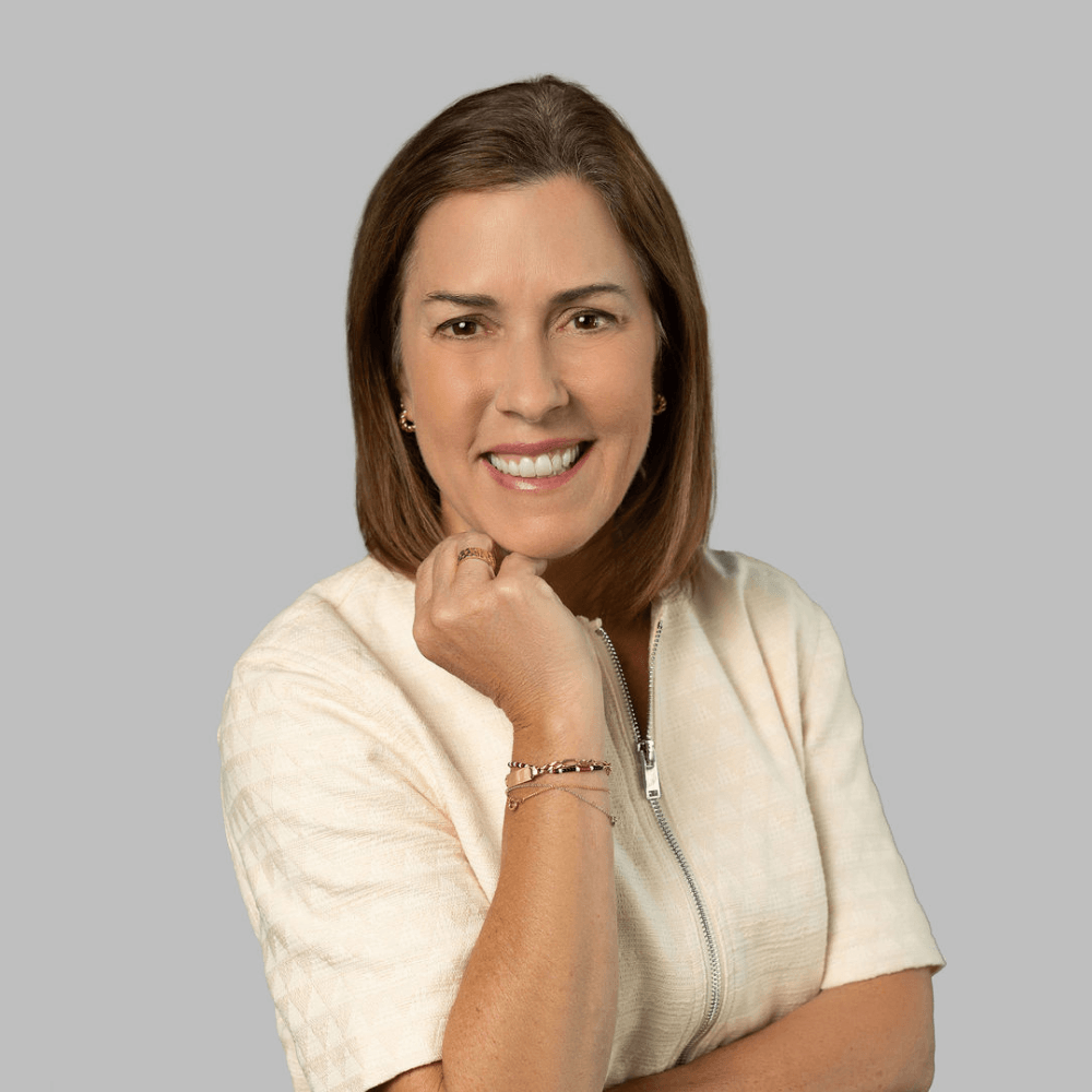 A woman with shoulder-length brown hair smiles while wearing a light-colored zip-up top, standing against a plain gray background. She has one hand raised to her chin and wears a bracelet on her wrist.