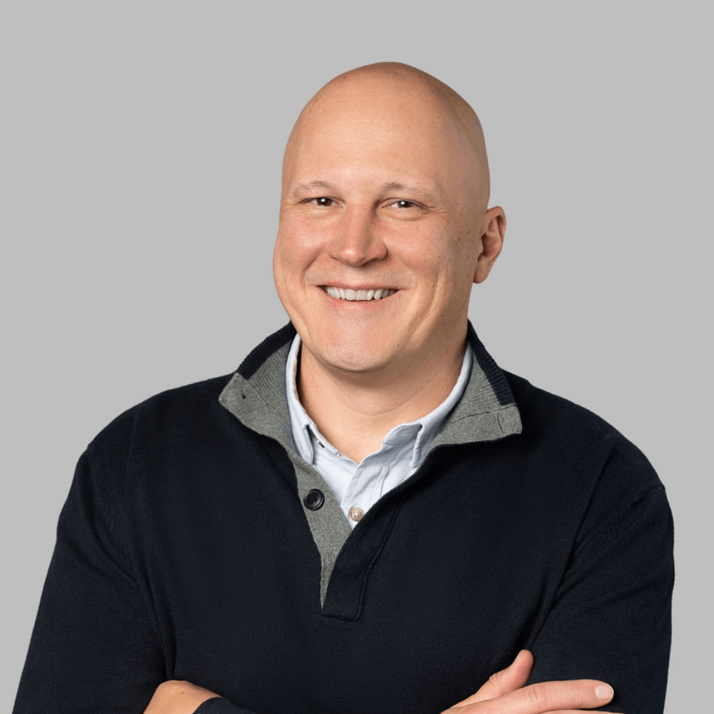 A smiling bald man wearing a dark pullover sweater over a collared shirt, standing with arms gently crossed in front of a plain light gray background.