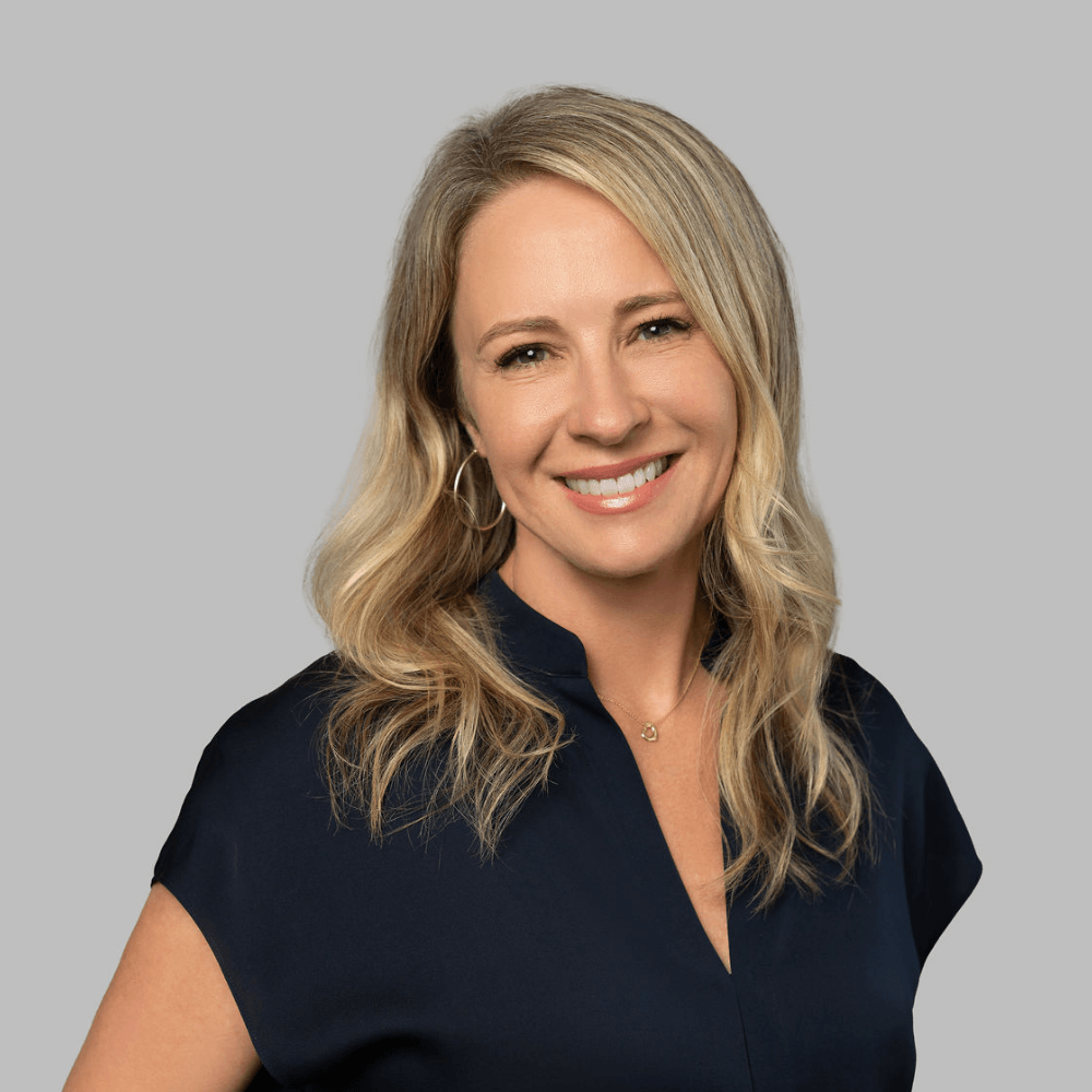 Smiling woman with wavy blonde hair, wearing a navy blouse and hoop earrings, posed in front of a plain light gray background.