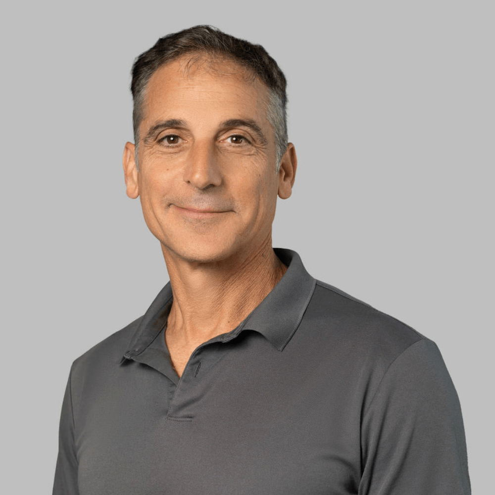 A middle-aged man with short brown and gray hair wearing a gray polo shirt, smiling softly, posed against a plain light gray background.
