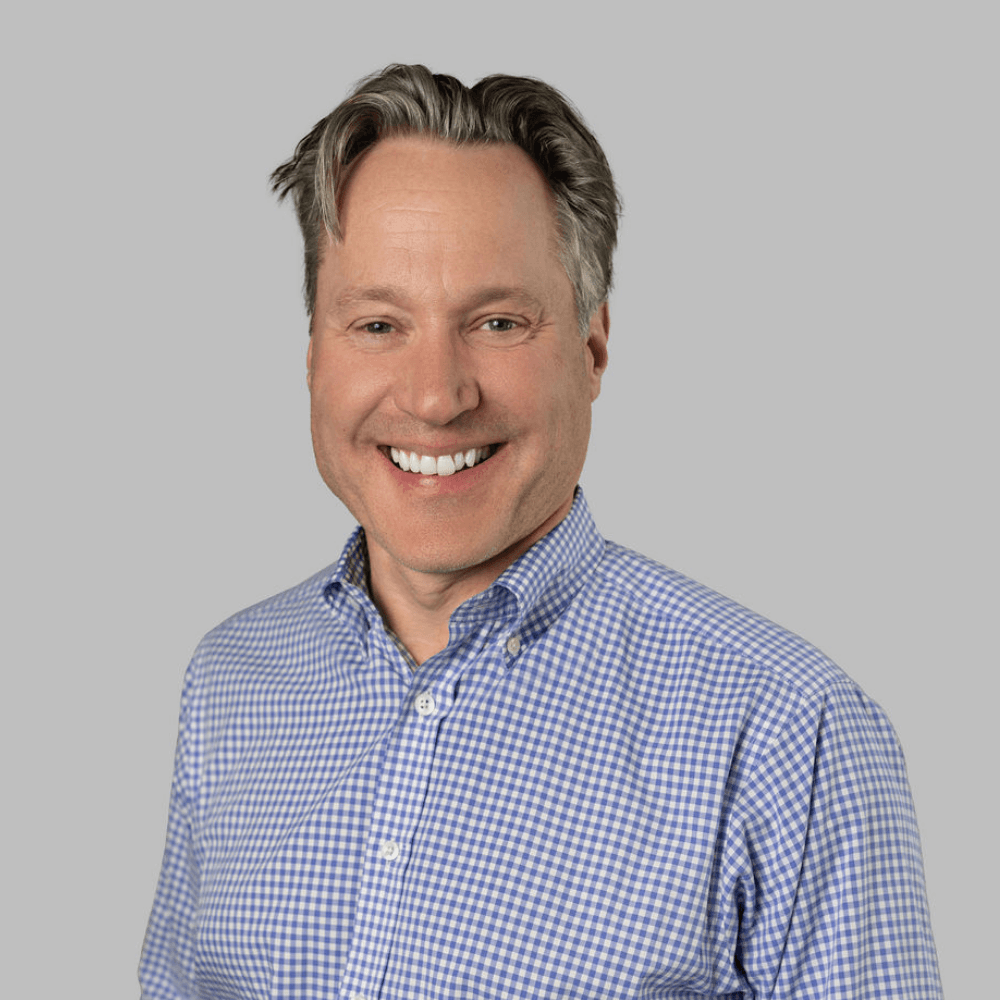 Smiling man with short gray hair wearing a blue and white checkered button-up shirt, posing in front of a plain light gray background.