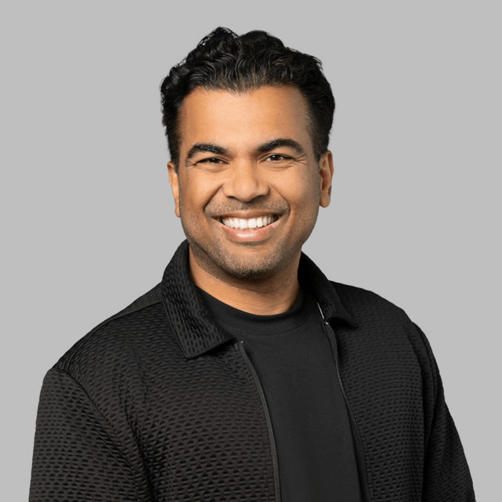 Smiling man with dark hair wearing a textured black jacket and black shirt, standing against a plain gray background.