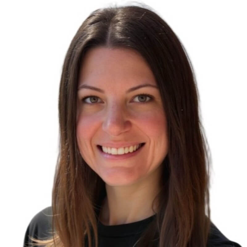 A woman with long, straight brown hair smiles at the camera. She is wearing a black shirt and is set against a plain white background, embodying the professionalism and approachability often seen among Outcomes Architects and CSMs.