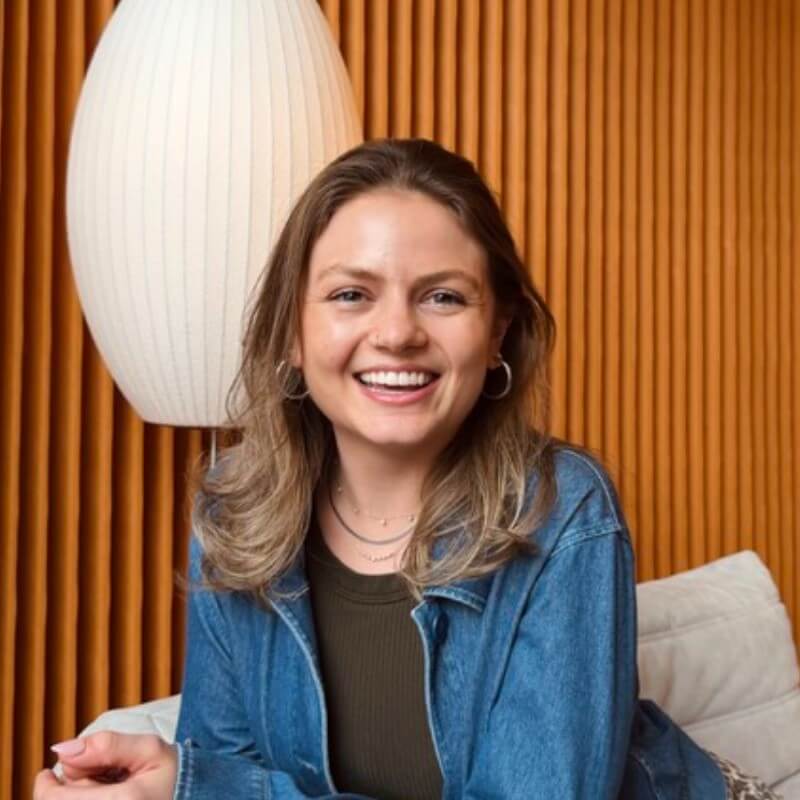 A smiling woman with light brown hair wearing a denim jacket sits on a couch in front of a wooden slat wall and a white pendant lamp, embodying the approachable spirit of Outcomes Architects and the collaborative energy that CSMs bring to every Notion workspace.