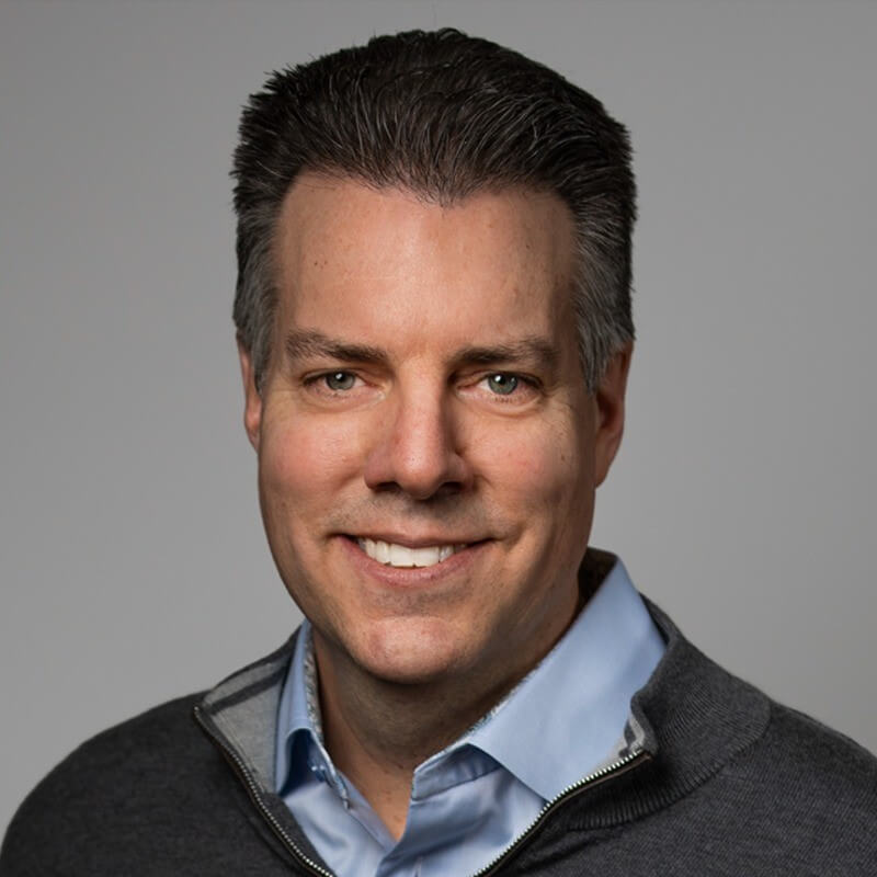 Headshot of a smiling man in a light blue collared shirt and grey quarter-zip sweater, an expert in churn reduction using Salesforce.