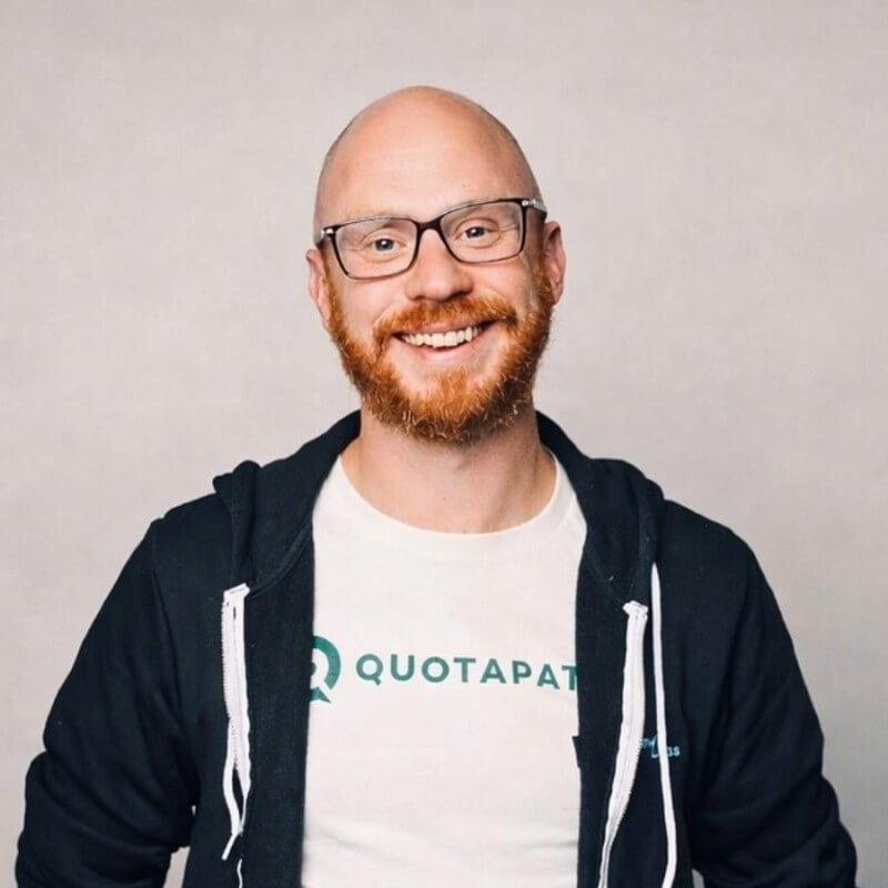 AJ Bruno, a smiling man with a red beard and glasses, wears a white QuotaPath T-shirt under a dark hoodie.
