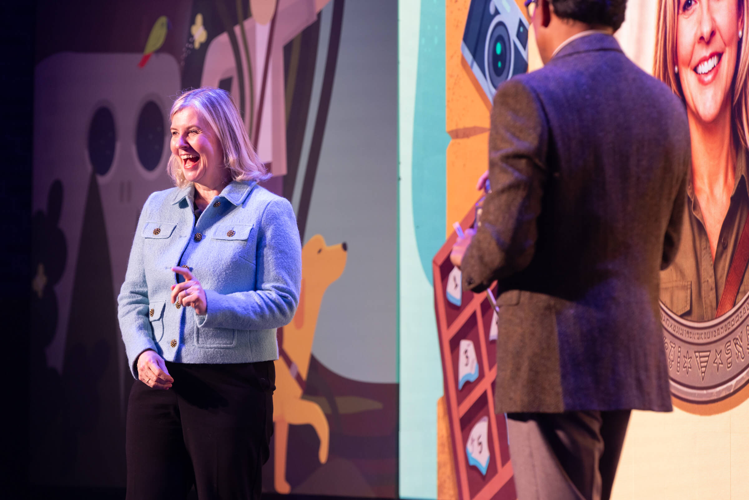A blonde woman in a light blue jacket laughs on stage during Pulse Europe 2026, with a man in a blazer standing with his back to the camera in the foreground.
