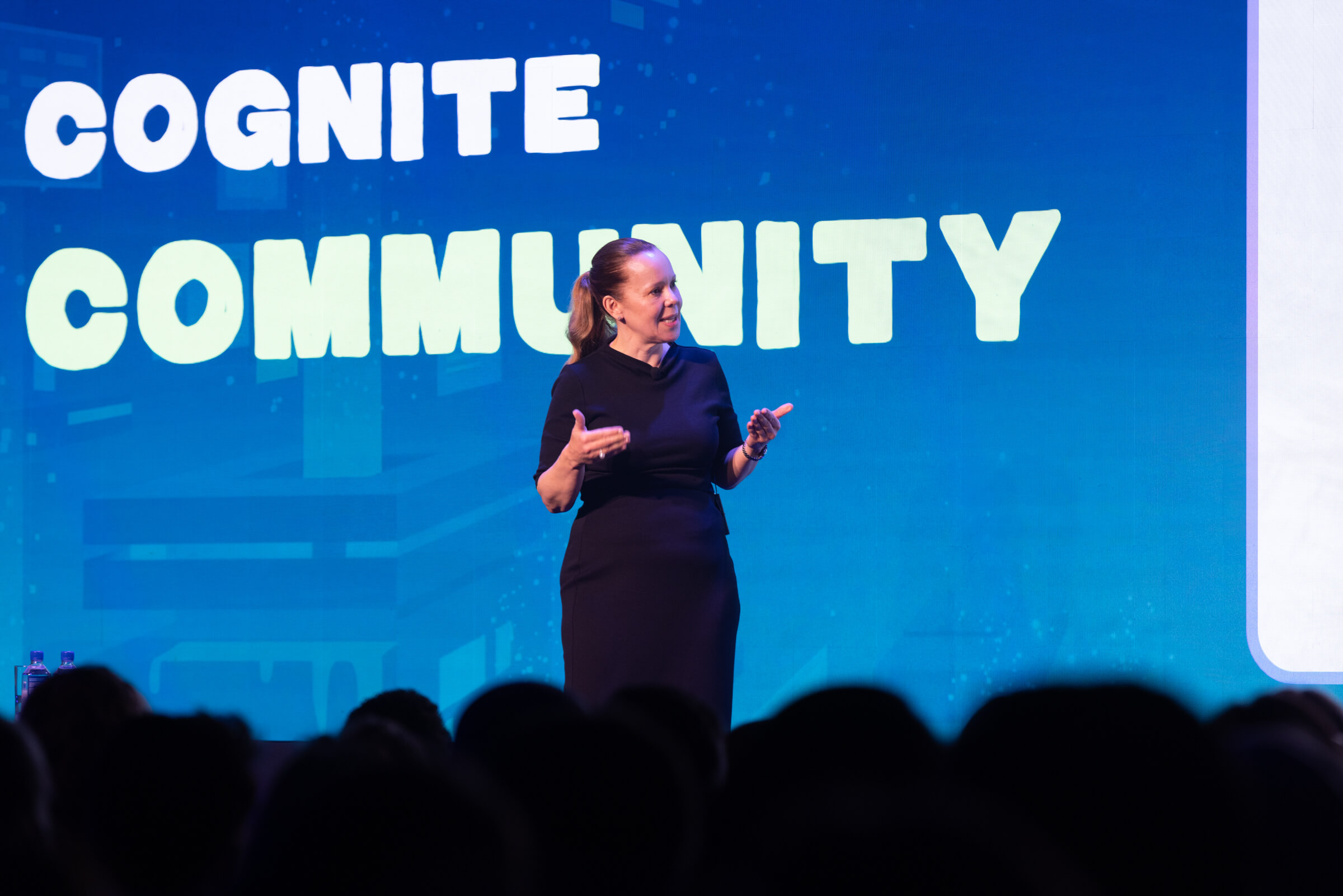A speaker stands on stage in front of a large blue screen promoting the Agentic Economy at Cognite Community.