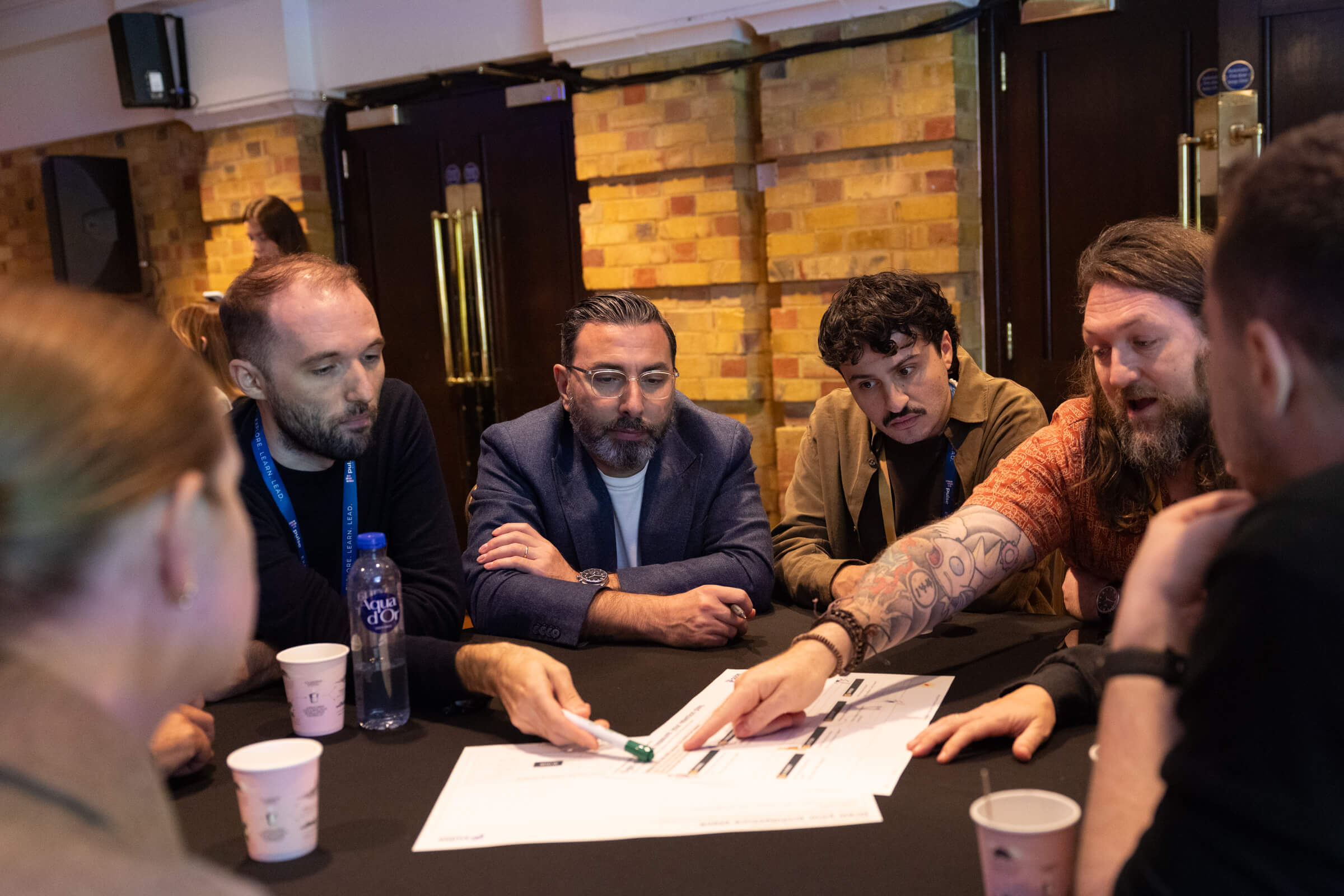 A group of five people sit around a black table in a brick-walled room, pointing to documents detailing retention strategies for the Agentic Economy.