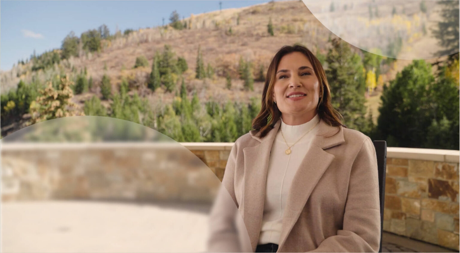 A woman in a beige coat sits on a patio with a mountain landscape in the background.